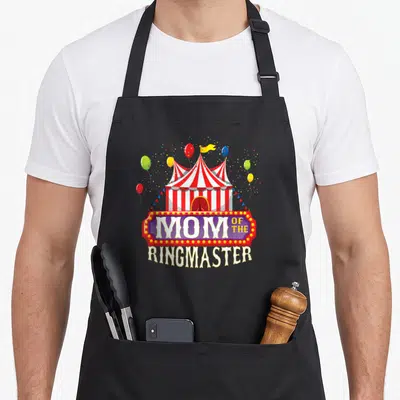 Ringmaster birthday apron - Circus tent with balloons and marquee sign reading Mom of the Ringmaster — shown on a black cotton canvas apron against a white background