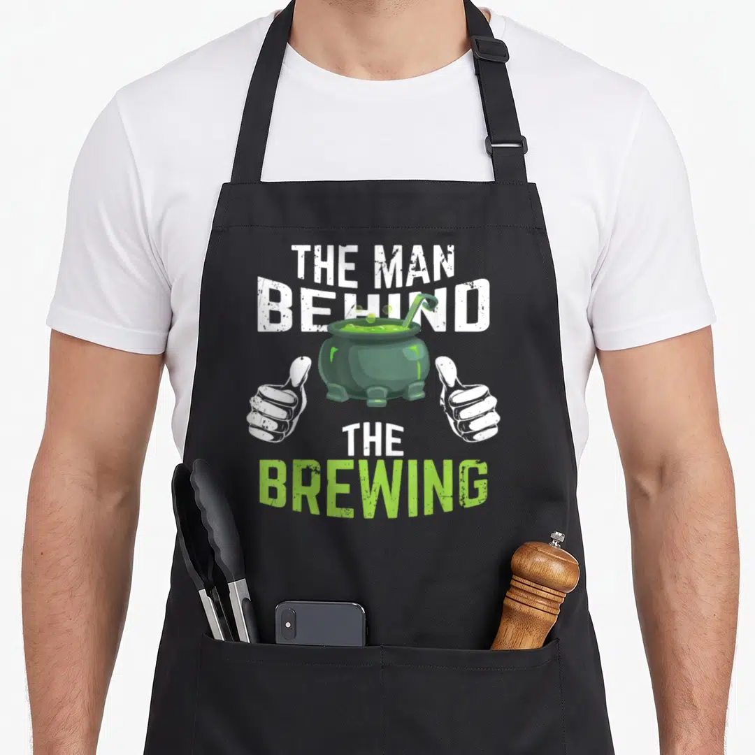 Behind halloween apron - Green cauldron between two white thumbs up hands with stacked text reading THE MAN BEHIND — shown on a black cotton canvas apron against a white background