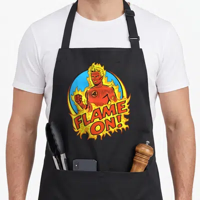 Fantastic flame apron - Retro Human Torch in blue circle with orange flames and FLAME ON! text — shown on a black cotton canvas apron against a white background