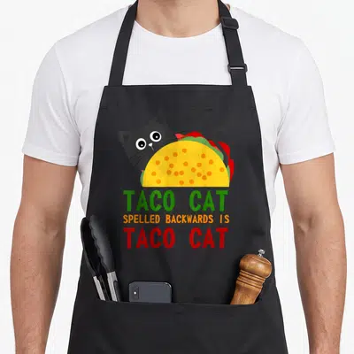 Cat taco apron - Cartoon black cat inside yellow taco with palindrome text TACO CAT — shown on a black cotton canvas apron against a white background