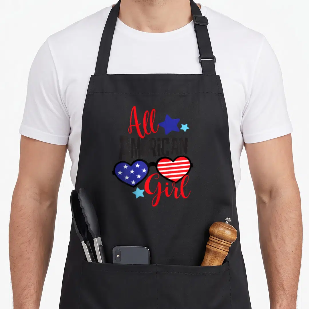 Funny patriotic 4th of july apron - All American Girl text with heart-shaped sunglasses stars stripes red white blue — shown on a black cotton canvas apron against a white background