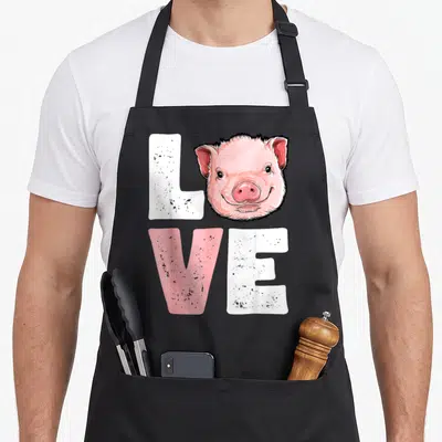 Pig farmers apron - Cartoon pig head replaces O in distressed LOVE text with pink and white letters — shown on a black cotton canvas apron against a white background