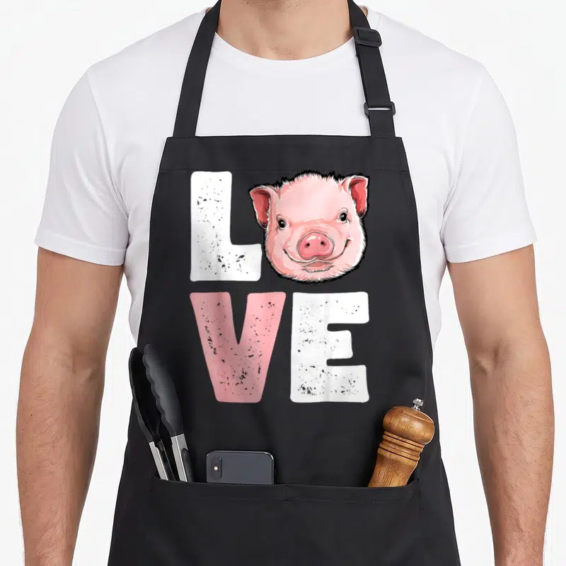 Pig farmers apron - Cartoon pig head replaces O in distressed LOVE text with pink and white letters — shown on a black cotton canvas apron against a white background