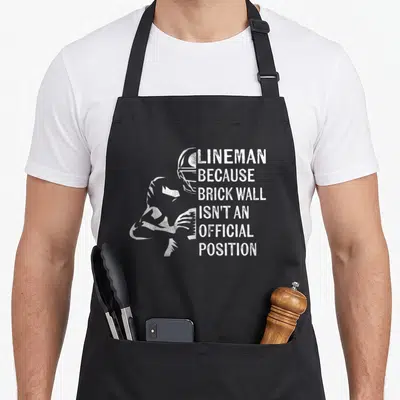 Funny lineman football apron - White distressed text reading Lineman Because Brick Wall next to football player — shown on a black cotton canvas apron against a white background