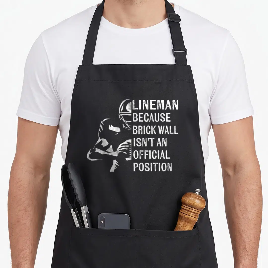 Funny lineman football apron - White distressed text reading Lineman Because Brick Wall next to football player — shown on a black cotton canvas apron against a white background