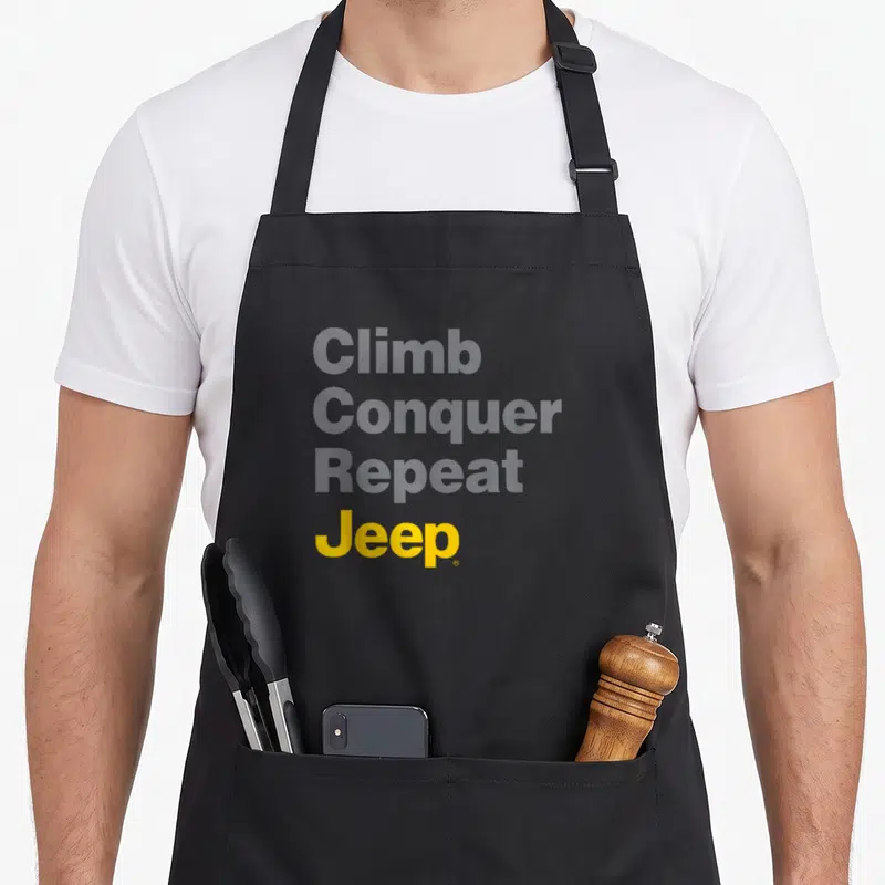 Climb jeep life apron - Stacked grey and yellow text reading Climb Conquer Repeat Jeep on a grey background — shown on a black cotton canvas apron against a white background