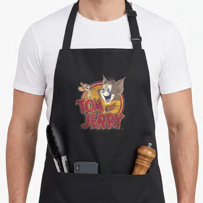 Water barbera apron - Tom and Jerry cartoon heads over red TOM and JERRY text on colorful circle — shown on a black cotton canvas apron against a white background