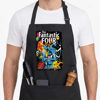 Epic first steps apron - The Fantastic Four comic cover with four heroes and descriptive text on paint splashes — shown on a black cotton canvas apron against a white background