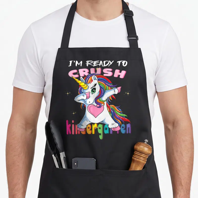 Ready unicorn aprons - Dabbing unicorn with rainbow mane and pink heart under I'm Ready To Crush kindergarten text — shown on a black cotton canvas apron against a white background