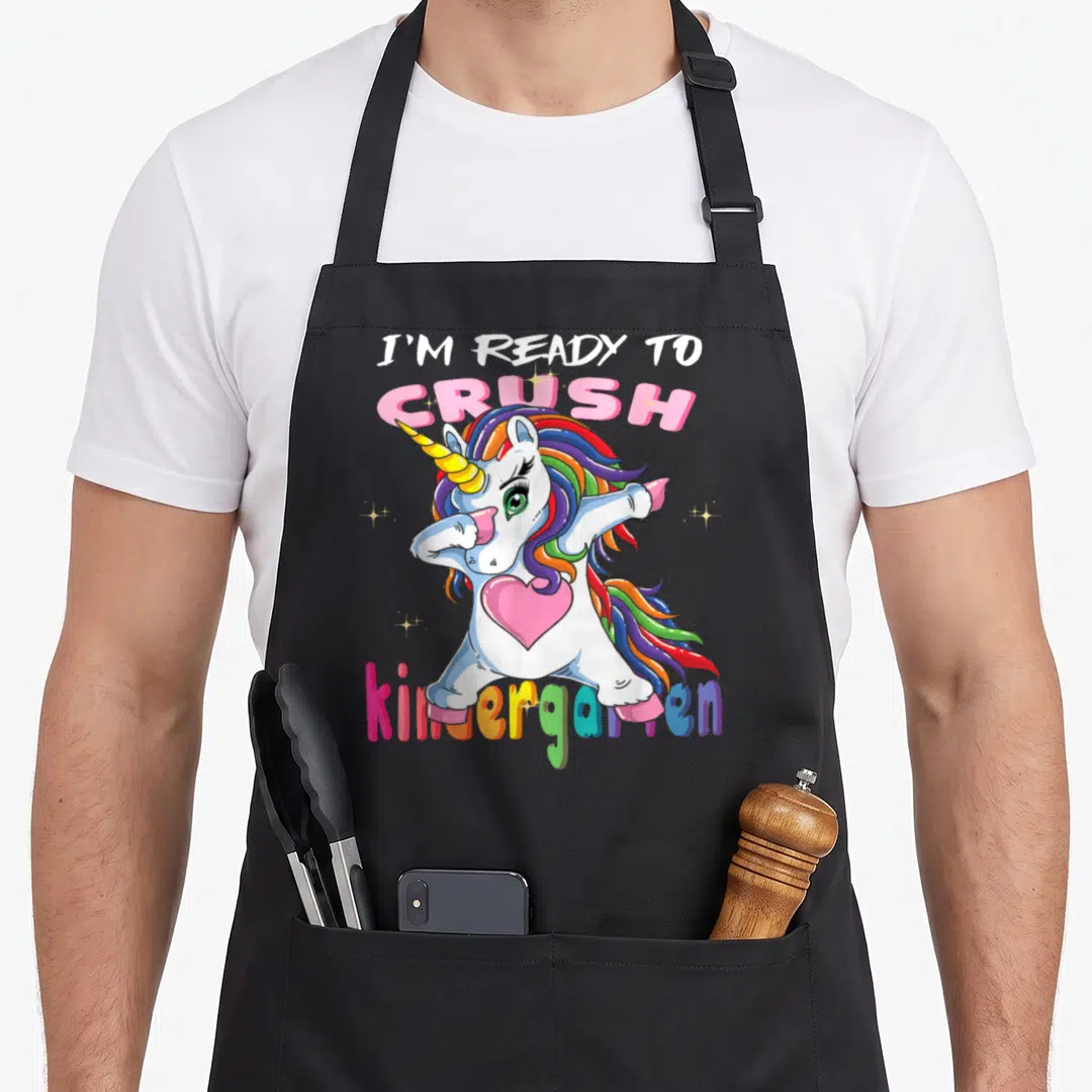 Ready unicorn aprons - Dabbing unicorn with rainbow mane and pink heart under I'm Ready To Crush kindergarten text — shown on a black cotton canvas apron against a white background