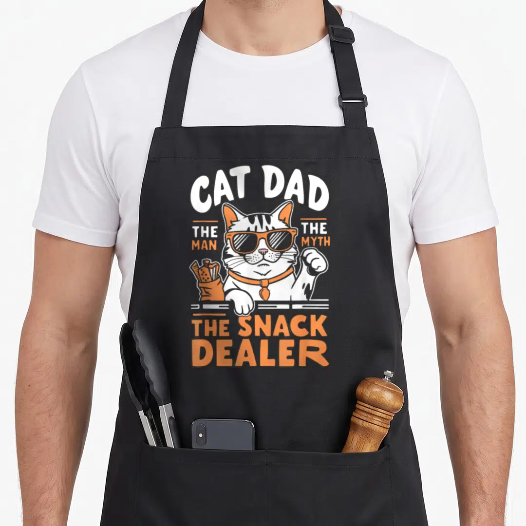 Myth cat dad apron - Orange and white cartoon cat with sunglasses holding a snack pouch surrounded by bold text — shown on a black cotton canvas apron against a white background
