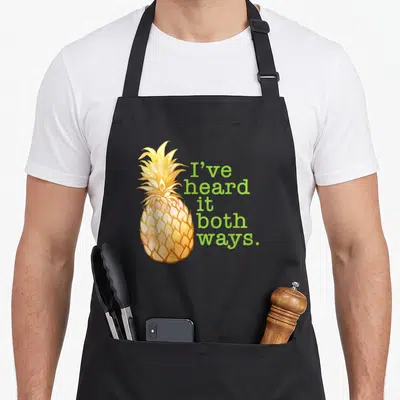 Heard pineapple apron - Watercolor yellow and orange pineapple next to green text reading I've heard it both ways — shown on a black cotton canvas apron against a white background