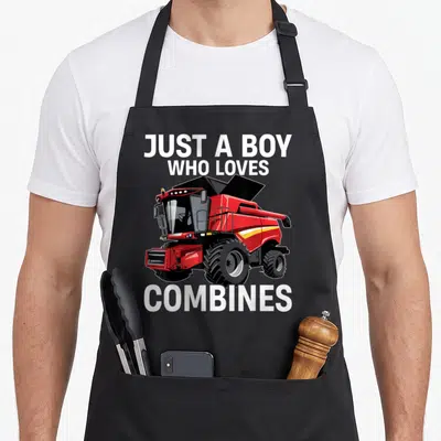 Funny harvester combine apron - Red combine harvester illustration with white text Just a Boy Who Loves Combines — shown on a black cotton canvas apron against a white background