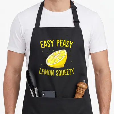 Easy lemonade apron - Yellow cartoon lemon slice with arched EASY PEASY LEMON SQUEEZY text and scattered white dots — shown on a black cotton canvas apron against a white background