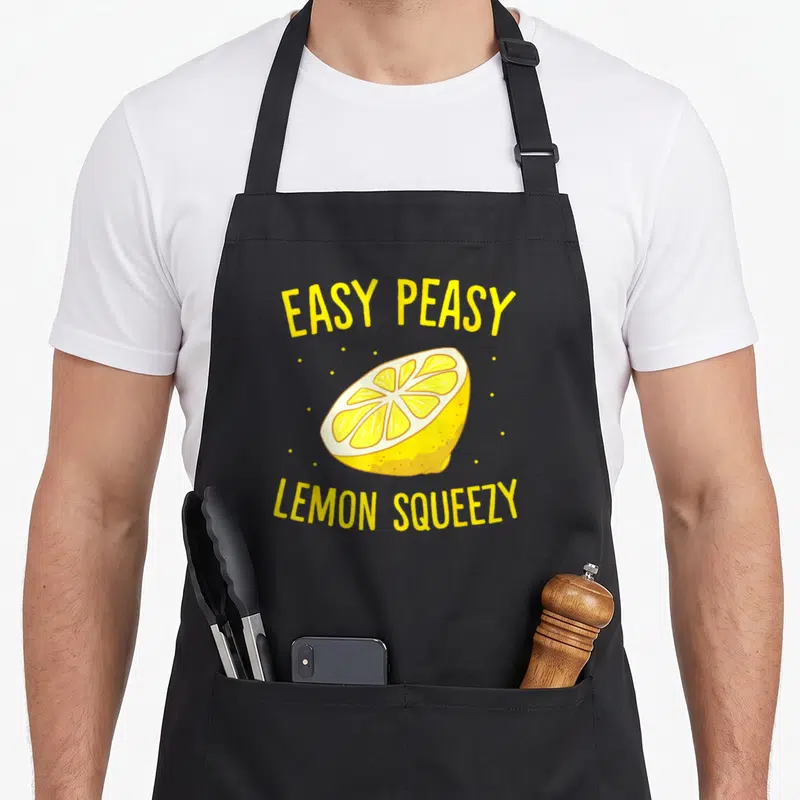Easy lemonade apron - Yellow cartoon lemon slice with arched EASY PEASY LEMON SQUEEZY text and scattered white dots — shown on a black cotton canvas apron against a white background
