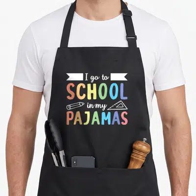 Pajamas teacher apron - Colorful text I go to SCHOOL in my PAJAMAS with pencil and ruler icons — shown on a black cotton canvas apron against a white background
