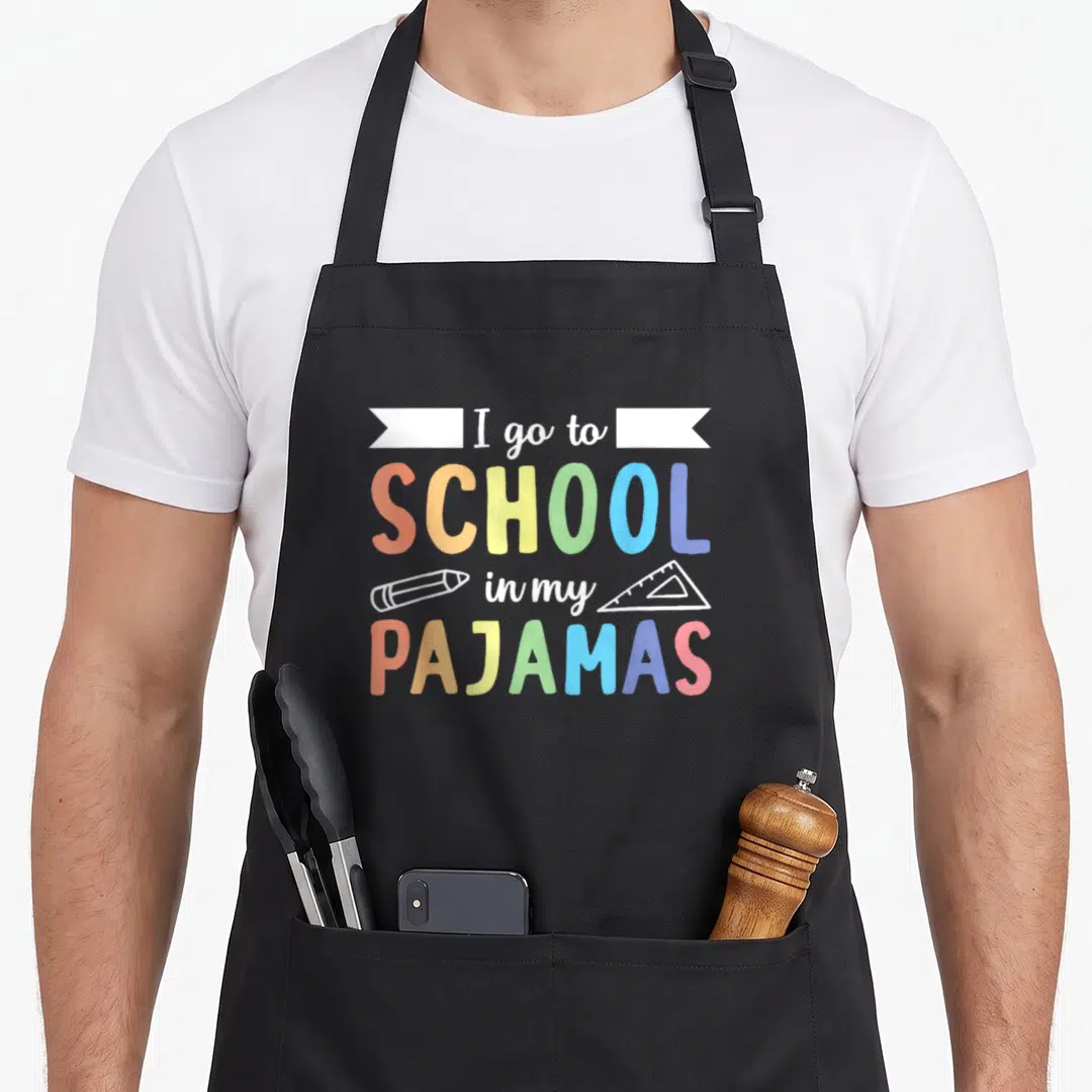 Pajamas teacher apron - Colorful text I go to SCHOOL in my PAJAMAS with pencil and ruler icons — shown on a black cotton canvas apron against a white background