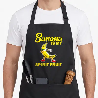Food banana apron - Cartoon banana character with green glasses and red shoes surrounded by yellow text reading Banana — shown on a black cotton canvas apron against a white background