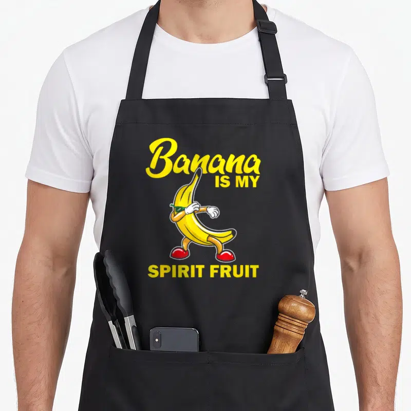 Food banana apron - Cartoon banana character with green glasses and red shoes surrounded by yellow text reading Banana — shown on a black cotton canvas apron against a white background