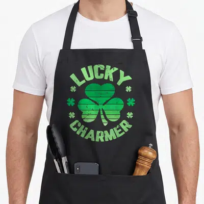 Lucky charmer st patrick day aprons - Green distressed shamrock with Lucky Charmer text in circular arrangement — shown on a black cotton canvas apron against a white background