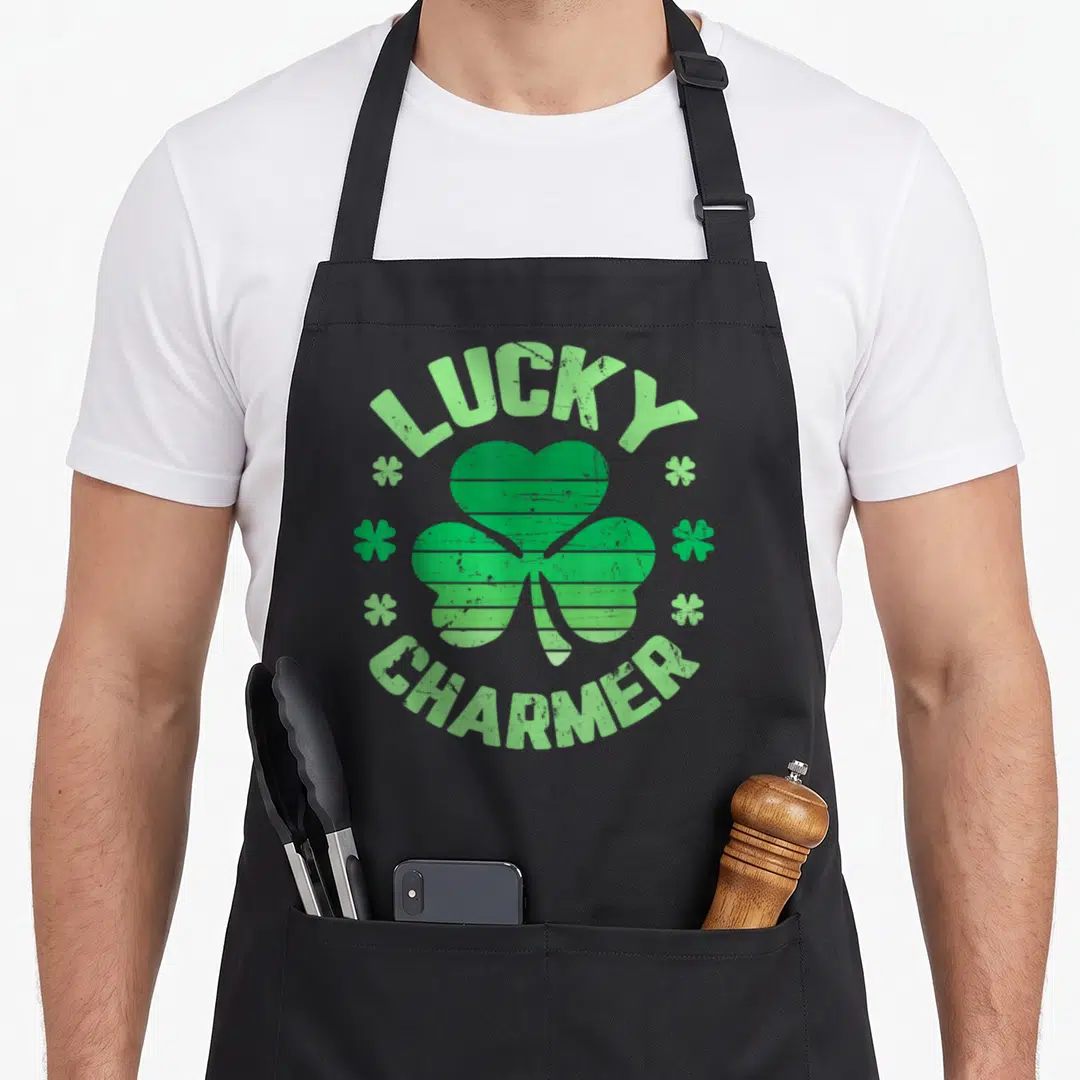 Lucky charmer st patrick day aprons - Green distressed shamrock with Lucky Charmer text in circular arrangement — shown on a black cotton canvas apron against a white background