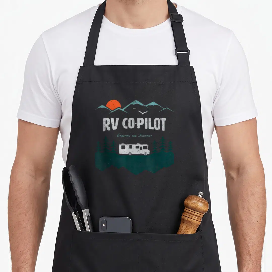 Pilot chef and co aprons - Retro RV Co-Pilot design with mountains, orange sun, trees, and white motorhome on green base — shown on a black cotton canvas apron against a white background