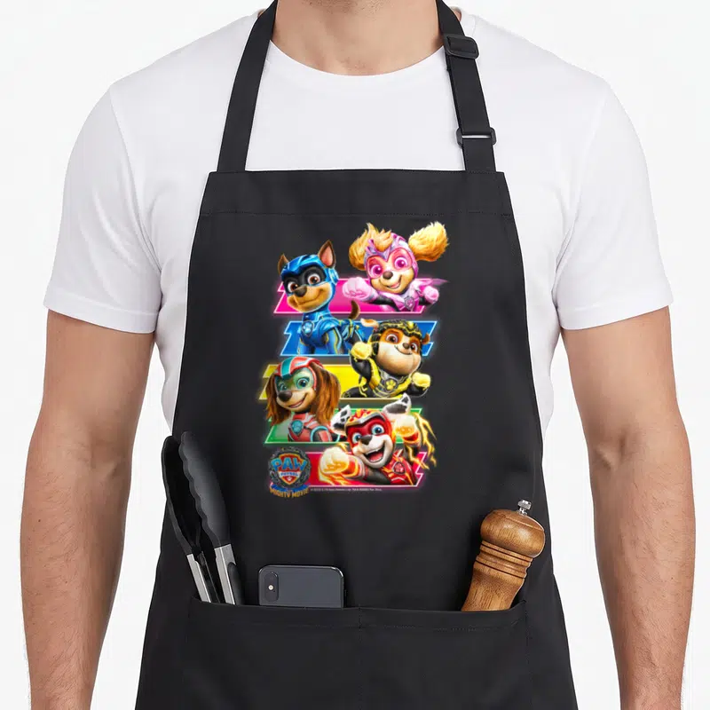 Mighty paw apron - Four puppies in superhero suits with PAW PATROL MIGHTY MOVIE text in blue red yellow and pink — shown on a black cotton canvas apron against a white background