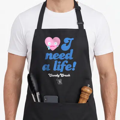 Funny cotton candy apron - Crying pink heart next to blue I need a life text and white Candy Crush logo — shown on a black cotton canvas apron against a white background