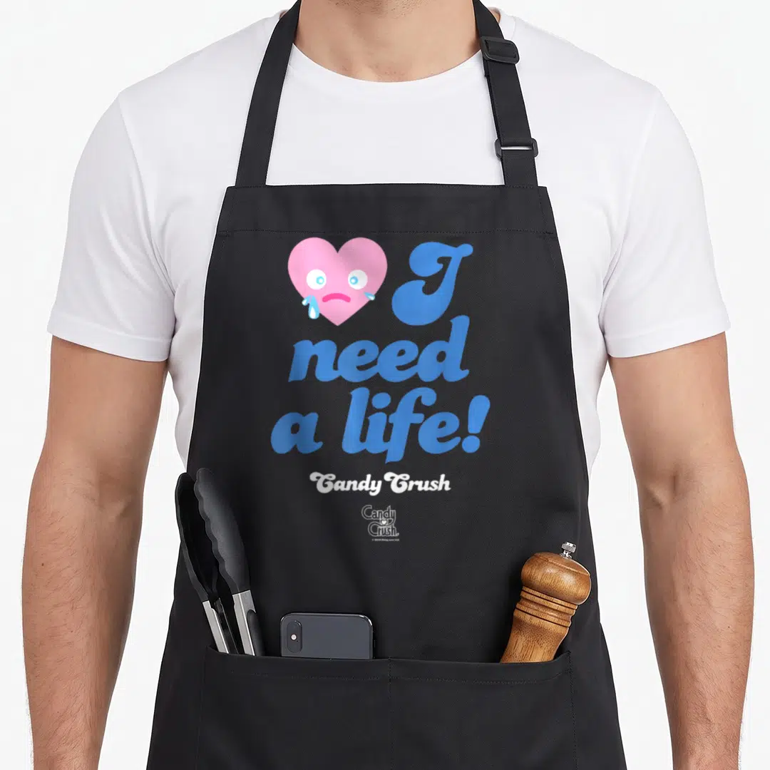 Funny cotton candy apron - Crying pink heart next to blue I need a life text and white Candy Crush logo — shown on a black cotton canvas apron against a white background