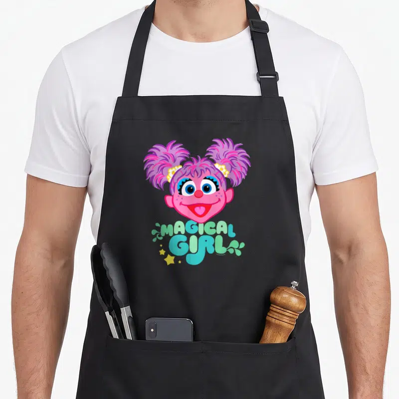 Abby street apron - Pink cartoon girl face with MAGICAL GIRL text surrounded by stars and leaves in pastel colors — shown on a black cotton canvas apron against a white background