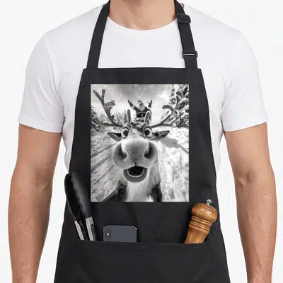 Selfie reindeer christmas apron - Black and white photo of a reindeer face close-up with Santa in a sleigh behind it — shown on a black cotton canvas apron against a white background