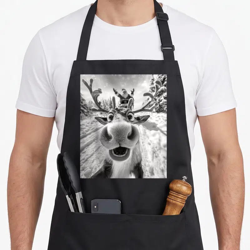 Selfie reindeer christmas apron - Black and white photo of a reindeer face close-up with Santa in a sleigh behind it — shown on a black cotton canvas apron against a white background