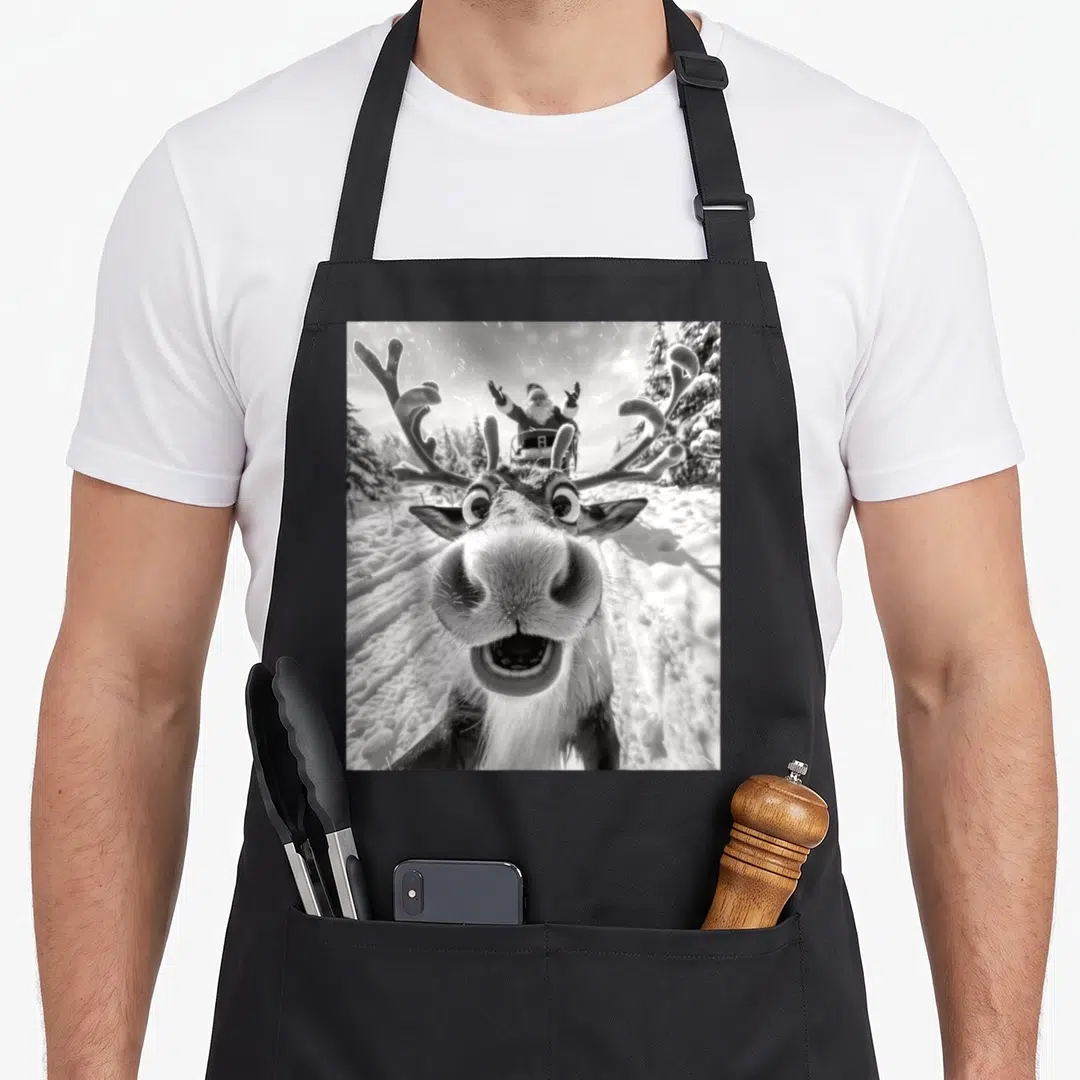 Selfie reindeer christmas apron - Black and white photo of a reindeer face close-up with Santa in a sleigh behind it — shown on a black cotton canvas apron against a white background