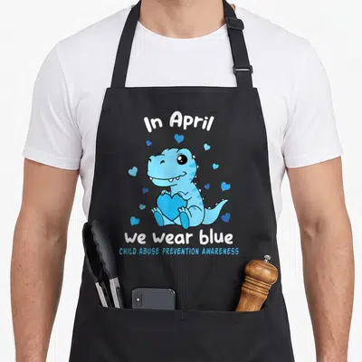 Dino blue april apron - Blue cartoon dinosaur holding heart surrounded by small hearts with awareness text — shown on a black cotton canvas apron against a white background