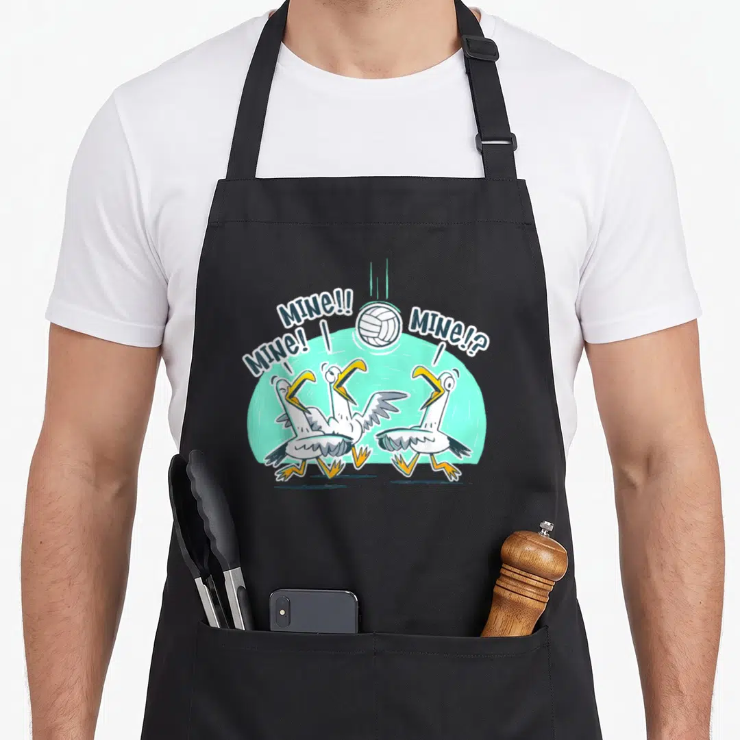 Volleyball bird apron - Three cartoon seagulls looking up at a volleyball with MINE speech bubbles — shown on a black cotton canvas apron against a white background