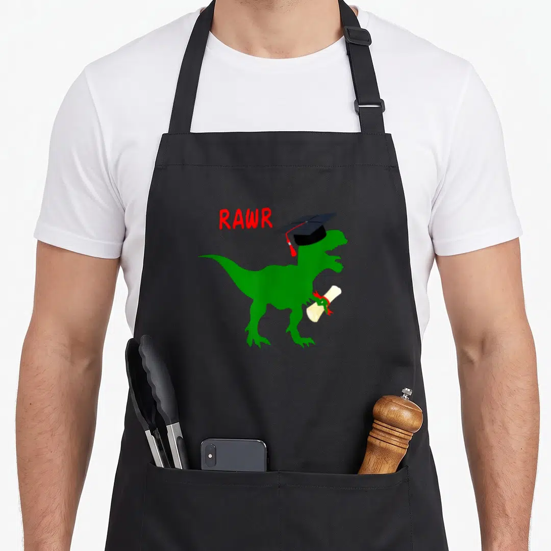 Diploma dinosaur apron - Green T-Rex wearing black grad cap holding beige diploma with red RAWR text — shown on a black cotton canvas apron against a white background