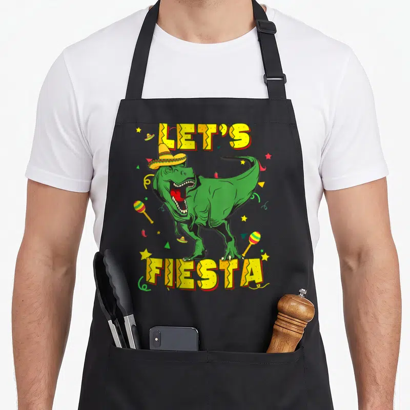 Cinco dinosaur apron - Green T-Rex in sombrero with LET'S FIESTA text surrounded by maracas chilies stars confetti — shown on a black cotton canvas apron against a white background
