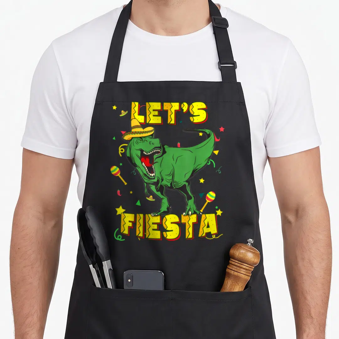 Cinco dinosaur apron - Green T-Rex in sombrero with LET'S FIESTA text surrounded by maracas chilies stars confetti — shown on a black cotton canvas apron against a white background