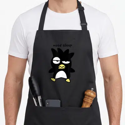 Badtz sleep deprived apron - Cartoon penguin in black white and yellow with text need sleep above it — shown on a black cotton canvas apron against a white background