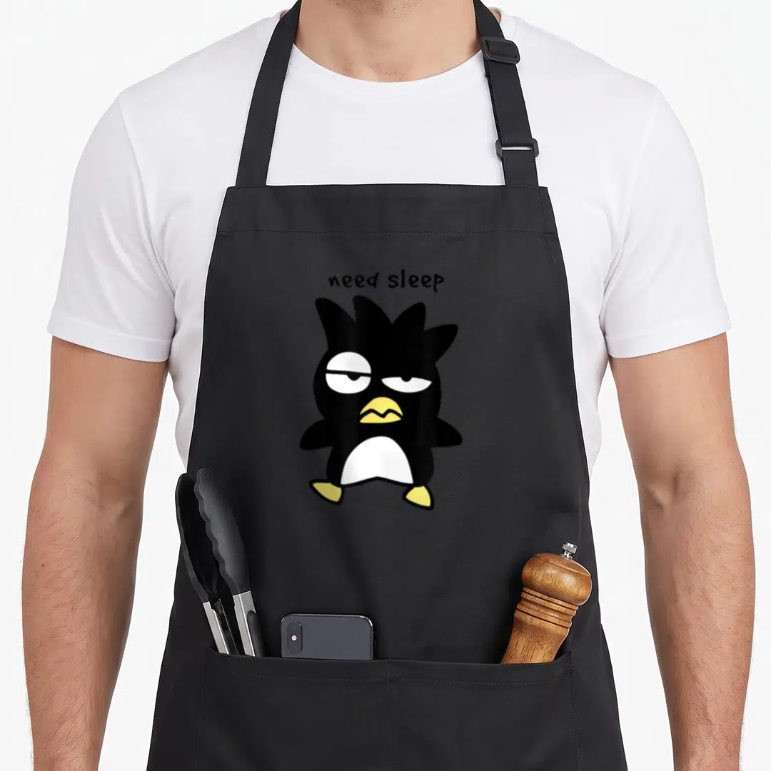 Badtz sleep deprived apron - Cartoon penguin in black white and yellow with text need sleep above it — shown on a black cotton canvas apron against a white background