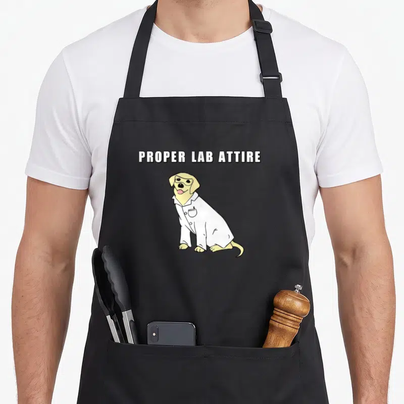 Proper laboratory apron - Cartoon yellow dog in white lab coat with text PROPER LAB ATTIRE — shown on a black cotton canvas apron against a white background