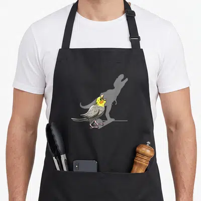 Shadow bird apron - Cartoon cockatiel with yellow head and grey body casting a dinosaur shadow — shown on a black cotton canvas apron against a white background