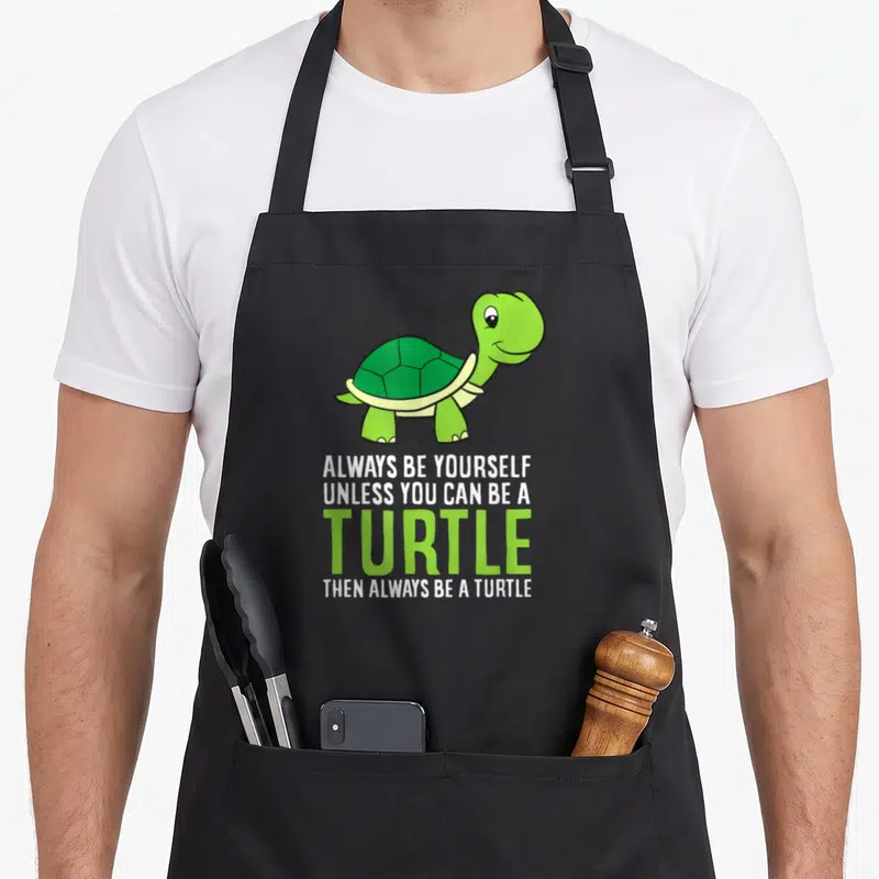 Sea pet apron - Cartoon green turtle above text Always Be Yourself Unless You Can Be A Turtle Then Always Be A Turtle — shown on a black cotton canvas apron against a white background