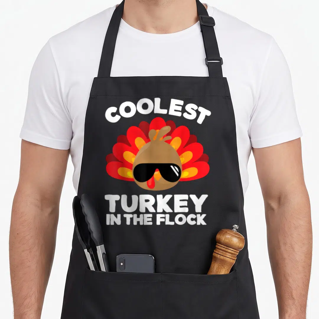 Coolest thanksgiving apron - Cartoon brown turkey head with black sunglasses and red feathers under white text — shown on a black cotton canvas apron against a white background