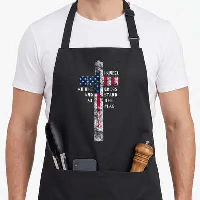 Kneel cross apron - Distressed cross filled with US flag pattern and split white text reading I kneel at the cross — shown on a black cotton canvas apron against a white background