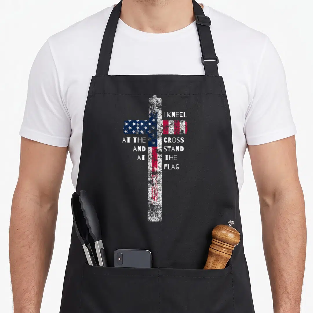 Kneel cross apron - Distressed cross filled with US flag pattern and split white text reading I kneel at the cross — shown on a black cotton canvas apron against a white background