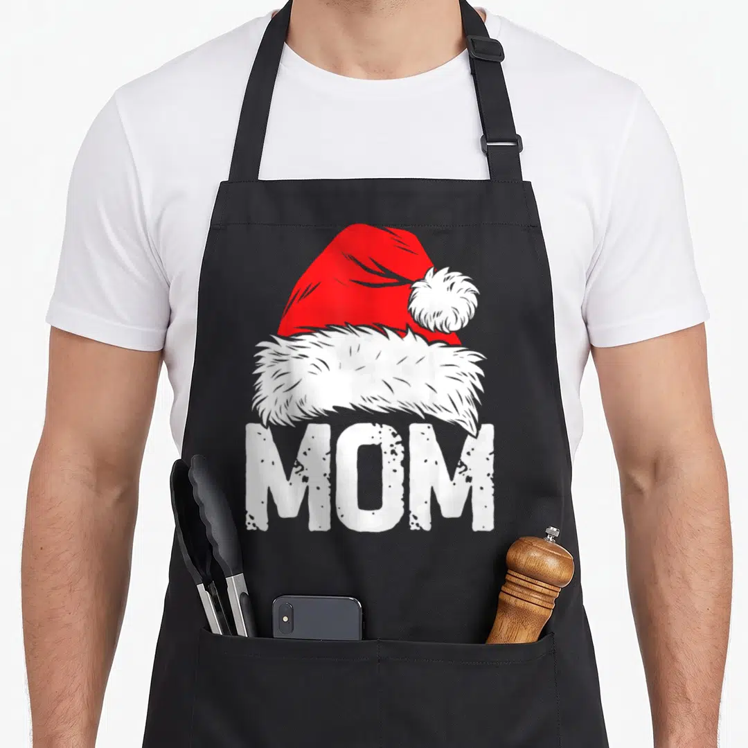 Santa mama apron - Red and white Santa hat above distressed white MOM text in cartoon style — shown on a black cotton canvas apron against a white background
