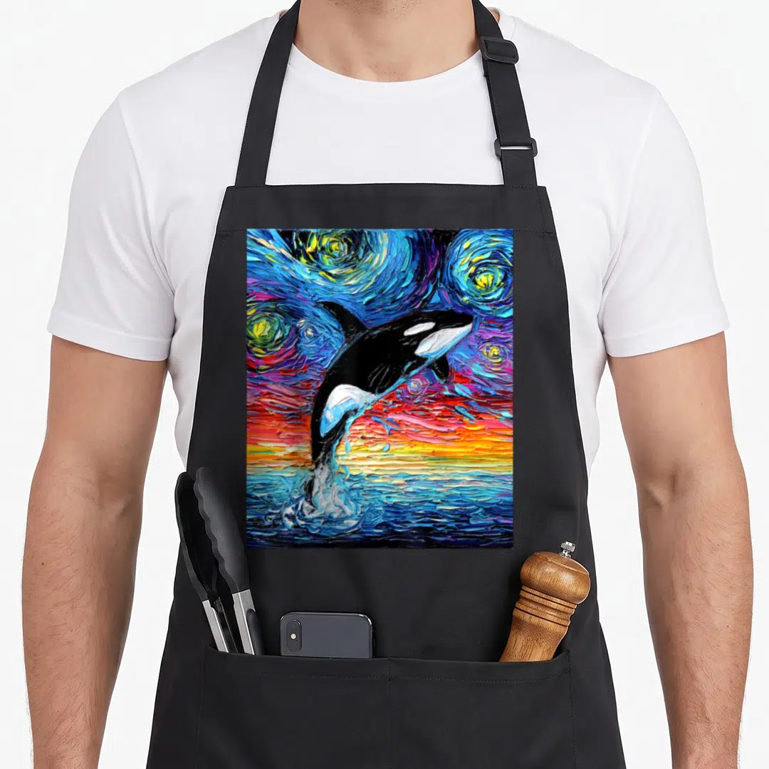 Orca colorful aprons - Black orca leaping from blue water into swirling starry sky with yellow and red strokes — shown on a black cotton canvas apron against a white background