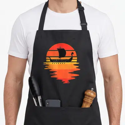 Water viking apron - Black viking ship silhouette on striped red and orange sunset with water reflection — shown on a black cotton canvas apron against a white background