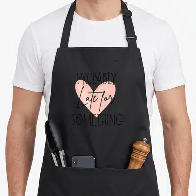 Probably girly apron - Pink heart with PROBABLY Late for SOMETHING text in black typography — shown on a black cotton canvas apron against a white background